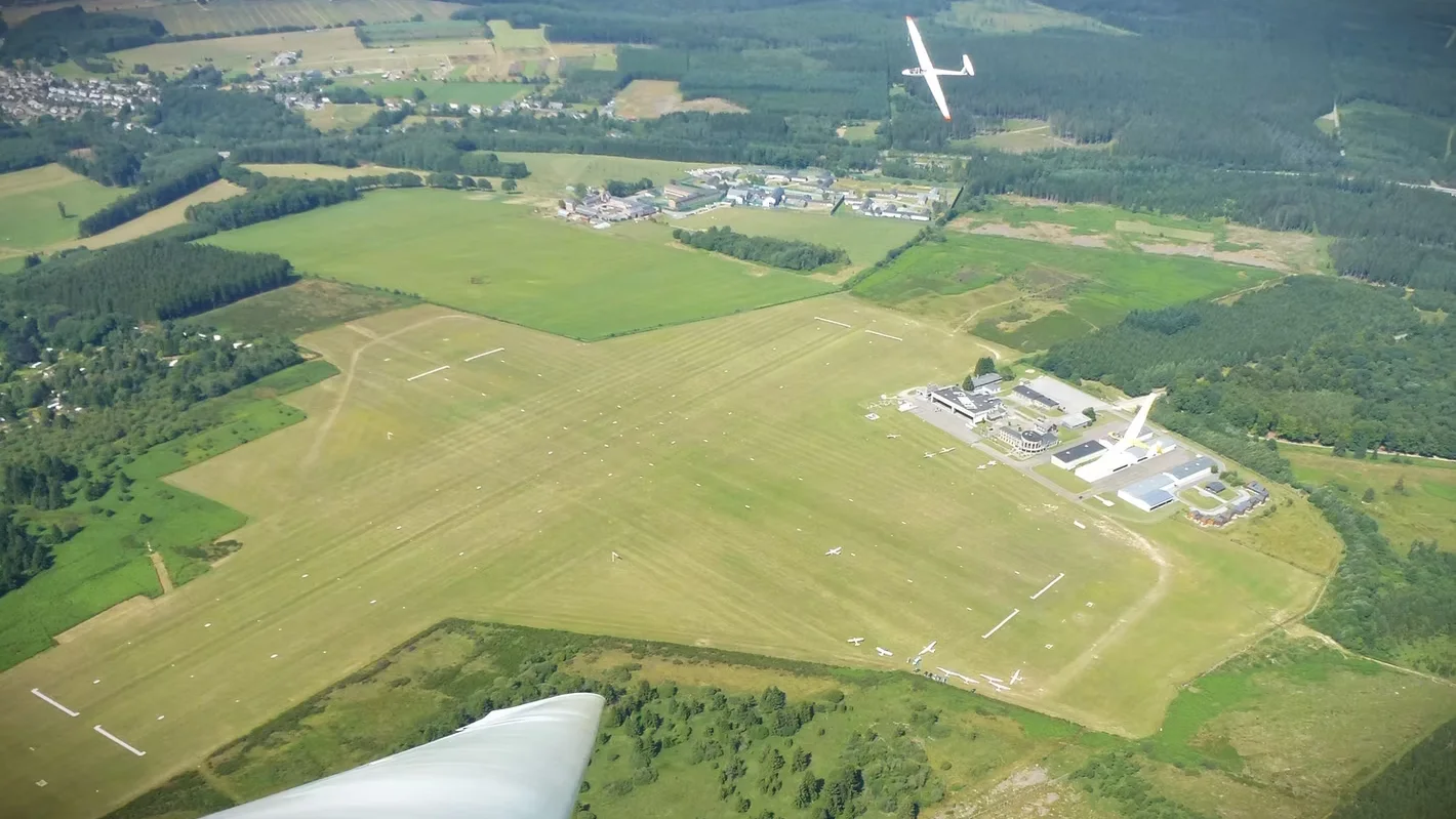 Vue aérienne de l’aérodrome de Saint-Hubert : deux pistes en herbe se croisant en X, aire de stationnement et hangars au nord-est, planeurs au sol et en vol.